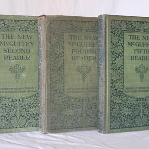 May include: Three antique hardcover books titled "The New McGuffey Second Reader," "Fourth Reader," and "Fifth Reader." The books have green covers with ornate floral designs and text in a rectangular frame. The publisher is the American Book Company.