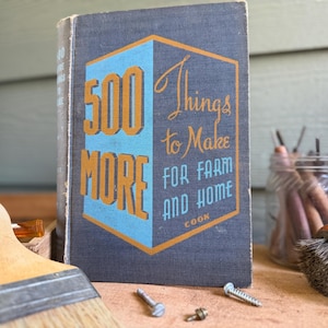 May include: Vintage book titled "500 More Things to Make for Farm and Home" with blue and orange lettering. A paintbrush, screws, and a jar of tools are in the foreground. The book's cover has a geometric design.