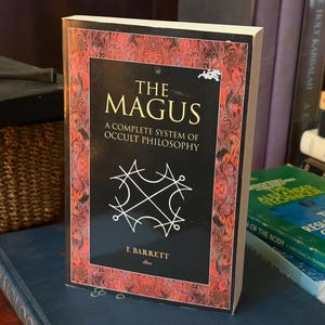 May include: A paperback book titled The Magus: A Complete System of Occult Philosophy by E. Barrett. The cover features a black central panel with white geometric line art, surrounded by a decorative red and black marbled border pattern.