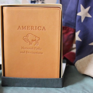 May include: A tan leather journal in a dark gray box. The journal is embossed with the word "AMERICA", a bison illustration, and "National Parks and Destinations". An American flag is in the background.