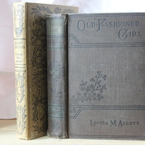 May include: Three antique books are displayed, with titles including "Eight Cousins" and "Old-Fashioned Girl" by Louisa May Alcott. The books have aged, textured covers in shades of tan and gray, with embossed floral and geometric designs.