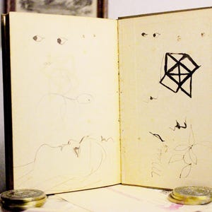 May include: An open book with sketches and drawings. The pages are off-white with brown ink drawings of eyes, faces, and geometric shapes. Two small, round, gold-colored containers are in the foreground.