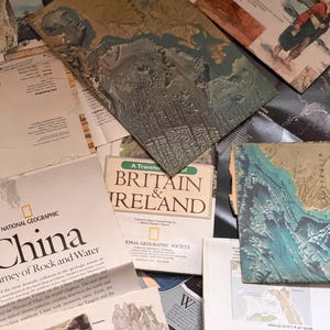 May include: A collection of National Geographic maps and travel guides featuring various regions including China, Britain, and Ireland. The assortment includes printed paper documents with topographical illustrations, geographic text, and historical imagery.