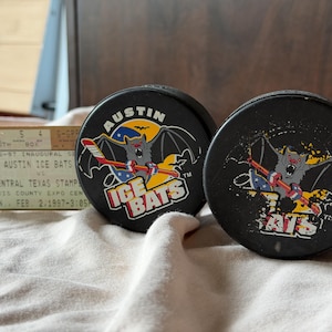 May include: Two black hockey pucks with the Austin Ice Bats logo, featuring a bat with a hockey stick. A vintage ticket stub for the 1996-97 Inaugural Season is visible in the background. The pucks are on a white cloth.