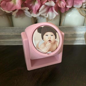 Op de afbeelding: Een roze fotolijst met een ronde foto van een baby met een bruine gebreide muts. De lijst is vierkant met een gebogen bovenkant en staat op een donkere houten ondergrond. Roze en witte bloemen staan op de achtergrond.