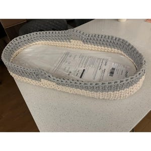 May include: A hand-crocheted baby bassinet in a soft, oval shape. The bassinet features alternating bands of light gray and cream-colored yarn. The interior is lined with a clear plastic sheet, likely for protection. A cozy and stylish nursery item.