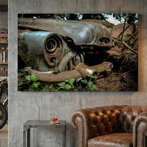 Könnte beinhalten: Leinwanddruck mit Nahaufnahme eines verwitterten Oldtimers. Das Bild zeigt das verrostete Metall, den Scheinwerfer und die Motorkomponenten, mit grünem Laub. Das Kunstwerk weckt Nostalgie und Automobilgeschichte.