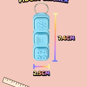 Może przedstawiać: Jasnoniebieski brelok typu fidget clicker z teksturowanymi przyciskami i trzema r&oacute;żnymi powierzchniami dotykowymi. Urządzenie ma 7,4 cm długości i 2,5 cm szerokości. Tekst na obrazku brzmi: TEXTURED BUTTON FIDGET CLICKER, 7.4CM, 2.5CM.