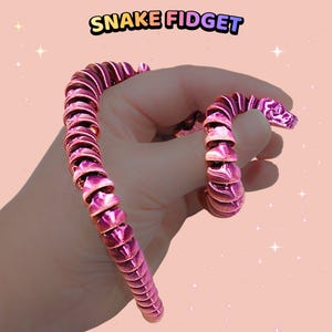 May include: A metallic magenta articulated snake fidget toy with a segmented, flexible body design. The text SNAKE FIDGET is printed in a colourful, rounded font at the top of the image against a light pink background with small white star accents.