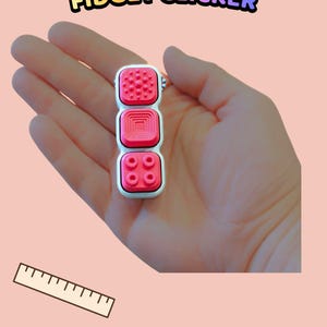Może przedstawiać: Prostokątne urządzenie typu fidget clicker z trzema r&oacute;żnymi r&oacute;żowymi przyciskami o zr&oacute;żnicowanych fakturach powierzchni, w tym wypukłymi kropkami, koncentrycznymi kwadratami i okrągłymi wypustkami, w białej ramce, z napisem TEXTURED BUTTON FIDGET CLICKER.