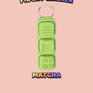 Może przedstawiać: Jasnozielony brelok typu fidget clicker z trzema kwadratowymi przyciskami, z kt&oacute;rych każdy ma inną fakturę powierzchni. G&oacute;rny przycisk ma małe wypukłe kropki, środkowy ma koncentryczne kwadraty, a dolny cztery koła. Tekst: TEXTURED BUTTON FIDGET CLICKER MATCHA.