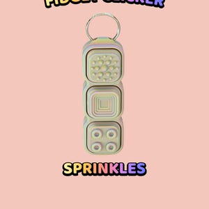 Może przedstawiać: Pastelowy brelok typu fidget clicker z trzema ułożonymi w stos kwadratowymi przyciskami o r&oacute;żnych wypukłych fakturach, w tym kropkami, kwadratem i k&oacute;łkami. Tekst "TEXTURED BUTTON FIDGET CLICKER" oraz "SPRINKLES" widnieje w kolorowych literach.