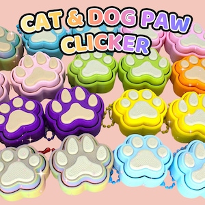 May include: A collection of paw-shaped clickers in various colours including purple, yellow, green, blue, pink, and orange. Each clicker features a textured beige pad design and is attached to a small beaded chain. The text CAT & DOG PAW CLICKER is visible.