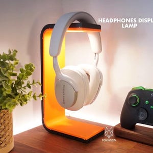 May include: A white headset is displayed on an orange and black headphone stand. The stand has a rectangular base and a curved back. The text "HEADPHONES DISPLAY LAMP" is visible. A black and green game controller is also present.