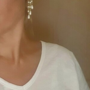 May include: A woman wearing a white v-neck t-shirt with a pair of dangling earrings. The earrings are gold with a cluster of clear rhinestones and a row of pearl-like drops.