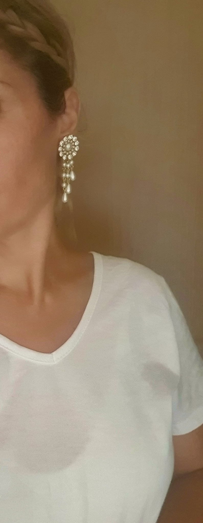 May include: A woman wearing a white v-neck t-shirt with a pair of dangling earrings. The earrings are gold with a cluster of clear rhinestones and a row of pearl-like drops.