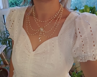 Op de afbeelding: Een witte, gelaagde parelketting met een gouden ketting en een bloemvormige hanger. De ketting wordt gedragen door iemand die een wit, kant-gezoomd topje draagt.