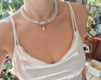 May include: A double-strand pearl choker necklace featuring a central silver-toned filigree clasp adorned with small clear crystals and a single dangling teardrop pearl pendant, worn with a white satin camisole top with thin spaghetti straps.