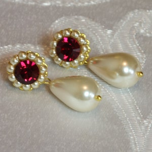 Puede incluir: Un par de pendientes de oro con una perla en forma de lágrima y un strass rojo rodeado de perlas blancas.