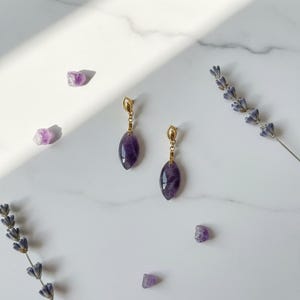 Peut inclure: Boucles d'oreilles pendantes en améthyste avec des détails dorés. Les boucles d'oreilles en forme de goutte sont présentées sur une surface en marbre blanc, avec des cristaux d'améthyste et des brins de lavande.