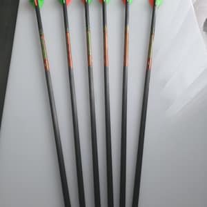 May include: Six black arrows with orange and green fletching are arranged on a white surface. The arrows have a dark shaft with orange and green accents. The fletching is a diamond shape.