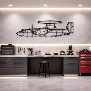 May include: Black metal wall art depicting a detailed outline of an aircraft with a distinctive radar dome. The artwork features a propeller and a tail fin, mounted on a light-colored wall in a workshop setting.
