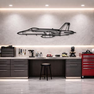 Pode incluir: Uma peça de arte de parede em metal preto que retrata um contorno detalhado de um caça. A obra de arte é montada em uma parede de cor clara acima de uma bancada com ferramentas e equipamentos. O jato apresenta asas, um cockpit e tanques de combustível externos.