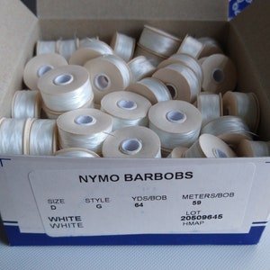 May include: A box of white Nymo Barbobs thread spools. The box is blue and white and has the text "Nymo Barbobs" printed on it. The spools are labelled "White" and have the following information: Size D, Style G, Yds/Bob 64, Meters/Bob 59, Lot 20509645, HMAP.