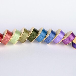 May include: A row of eight spools of thread in various colors, including red, green, blue, and purple. The spools are arranged in a diagonal line, with the red spool at the bottom left and the purple spool at the top right.