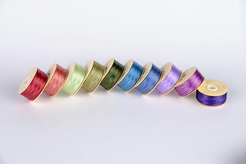 May include: A row of eight spools of thread in various colors, including red, green, blue, and purple. The spools are arranged in a diagonal line, with the red spool at the bottom left and the purple spool at the top right.