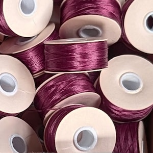 May include: Close-up of spools of deep burgundy thread wound around beige cardboard bobbins. The thread is smooth and shiny.