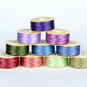 Può includere: Una collezione di dieci bobine di filo di vari colori, tra cui viola, blu, verde e rosso. Le bobine sono disposte a forma di piramide.