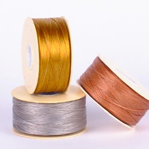 May include: Three spools of thread, one gold, one brown, and one gray, are stacked on top of each other.