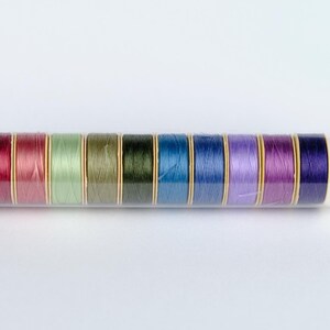 May include: A clear plastic container holds 10 spools of thread. The spools are arranged in a row and are in various colors, including red, green, blue, and purple.