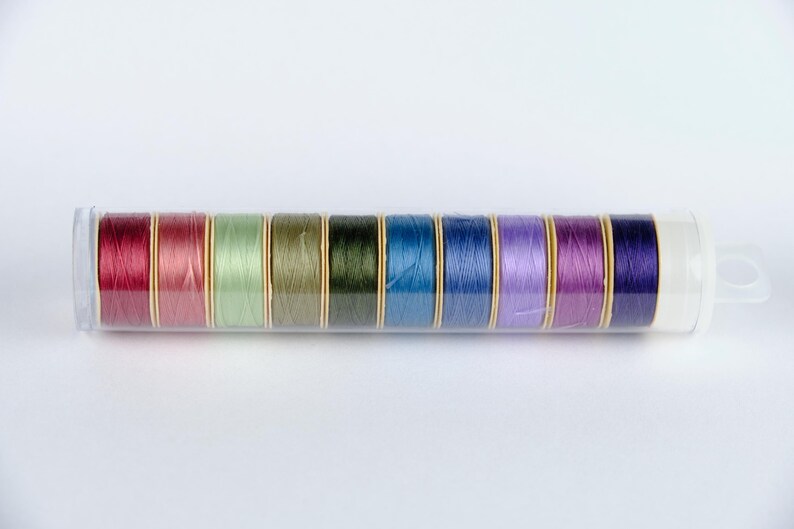 May include: A clear plastic container holds 10 spools of thread. The spools are arranged in a row and are in various colors, including red, green, blue, and purple.