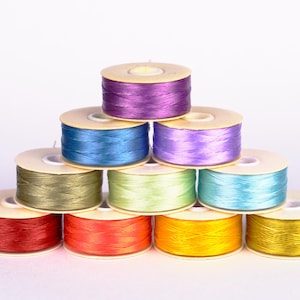 May include: A pyramid of colorful thread spools in shades of red, orange, yellow, green, blue, and purple. The spools are arranged on a white background, showcasing the vibrant colors of the thread. Ideal for sewing and crafting projects.