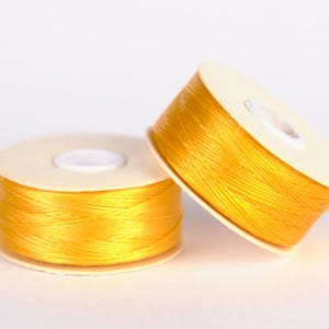 Puede incluir: Dos carretes de hilo amarillo, enrollados en bobinas de plástico blanco.