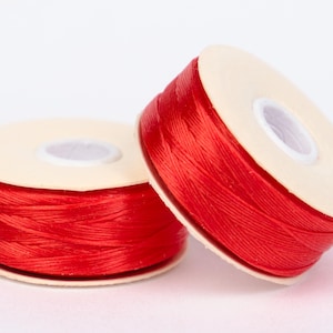 Puede incluir: Dos carretes de hilo rojo brillante. El hilo está enrollado firmemente alrededor de un núcleo de cartón beige. Los carretes están colocados sobre una superficie blanca, uno ligeramente inclinado contra el otro. El hilo parece ser un material liso y brillante.