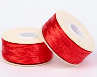 May include: Two spools of bright red thread. The thread is tightly wound around a beige cardboard core. The spools are positioned on a white surface, with one slightly leaning against the other. The thread appears to be a smooth, shiny material.