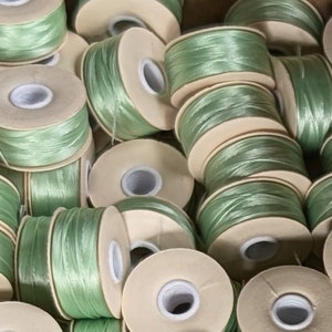May include: A close-up of a pile of spools of pale green thread. The spools are made of cardboard and have a white center.