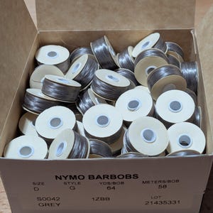 May include: A cardboard box filled with multiple grey Nymo BarBobs thread bobbins. The box is labeled with text including NYMO BARBOBS, SIZE D, STYLE G, 64 YDS/BOB, 58 METERS/BOB, S0042 GREY, 1ZBB, and LOT 21435331. Each bobbin has a white core.