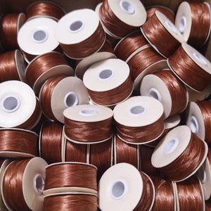 May include: A close-up of a pile of brown thread spools. The spools are made of cardboard and have a white center. The thread is wound around the spools and is a rich brown color.