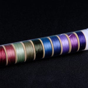 May include: A clear plastic container filled with ten spools of thread. The spools are arranged in a row and are in various colors, including red, green, blue, and purple.