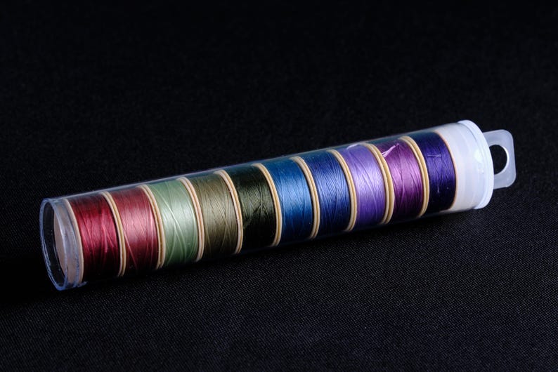 May include: A clear plastic container filled with ten spools of thread. The spools are arranged in a row and are in various colors, including red, green, blue, and purple.