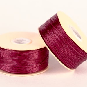 May include: Two spools of burgundy thread wound on beige plastic bobbins.