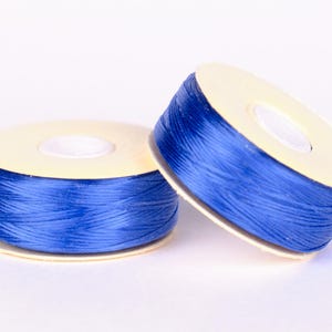 May include: Two spools of blue thread. The thread is tightly wound around a light tan cardboard core. The spools are positioned on a white background. The thread appears to be a vibrant shade of blue.