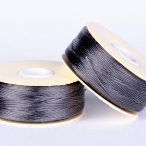 May include: Two spools of black thread are displayed against a white background. The thread is tightly wound around a light tan cardboard core. The spools are positioned at different angles, showcasing the thread's texture and color.