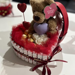 May include: A heart-shaped gift basket with a brown teddy bear holding a purple heart. The basket is filled with chocolates, white roses, and red tulle. Two red glitter heart decorations are attached to the basket.