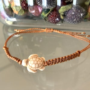 Può includere: Un braccialetto in macramè marrone con un ciondolo a forma di tartaruga color crema. Il braccialetto ha una chiusura scorrevole regolabile. Il ciondolo a forma di tartaruga è centrato e presenta dettagli sul guscio.