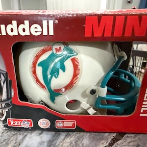 Könnte beinhalten: Ein Riddell Mini-Footballhelm in einer rot-weißen Schachtel. Der weiße Helm zeigt ein türkisfarbenes Delfin-Logo mit rotem Sonnenstrahlen-Motiv und dem Buchstaben M. Der Helm hat einen türkisfarbenen Gesichtsschutz. Die Box zeigt Riddell, MINI und FOOTBALL HELMET.